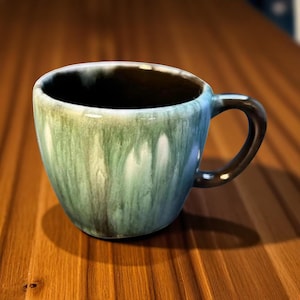 以下が含まれることがあります： ユニークな釉薬を施したセラミックコーヒーマグ。マグカップは、緑、青、茶色の色のブレンドで、濃い茶色のハンドルが付いています。内側は濃い茶色です。マグカップは丸みを帯びた形状です。