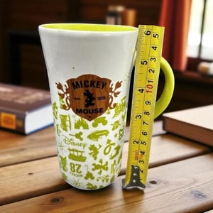 May include: White ceramic Mickey Mouse mug with a yellow interior and handle. The mug features a brown and green Mickey Mouse logo and various green Disney-themed icons. The mug is approximately 5 inches tall.
