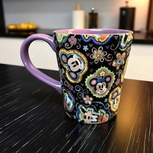 以下が含まれることがあります： 紫色の持ち手と縁が付いたセラミックマグ。黒い背景にカラフルなミッキーマウスとミニーマウスのデザインが施されています。マグカップは鮮やかで模様のある美しさを持っています。