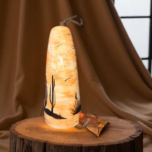 Peut inclure: Une lampe cylindrique avec un motif de paysage désertique. La lampe présente une palette de couleurs chaudes, orange et crème, avec des silhouettes de cactus noires. Elle possède une poignée en corde et repose sur une base en bois. La lampe est éclairée de l'intérieur.
