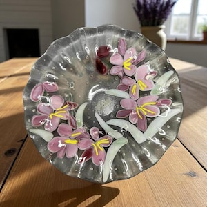 May include: A decorative glass dish with a scalloped edge. The dish is clear with embedded pink flowers, yellow stamens, and green leaves. The flowers are clustered in the center of the dish. The dish is on a wooden surface.