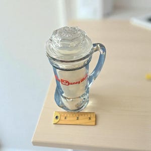 May include: Clear glass mug with a handle and lid. The mug has the words "Disney World" in red and white. A wooden ruler is next to the mug. The mug is approximately 3 inches tall.