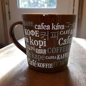 May include: A brown ceramic coffee mug with a curved handle. The mug is printed with the word "coffee" in multiple languages, including English, Korean, and Japanese. The mug is sitting on a wooden surface.