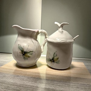 Puede incluir: Un juego de crema y azucarero de cerámica color crema. Cada pieza presenta un diseño de mariposa pintado a mano en tonos verdes y amarillos. El azucarero tiene tapa y cucharita.