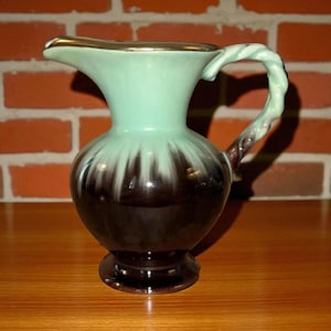 May include: A vintage ceramic pitcher with a mint green upper half and a dark brown lower half. The pitcher has a gold rim and a twisted handle. The design features a drip effect where the green paint transitions to the brown.