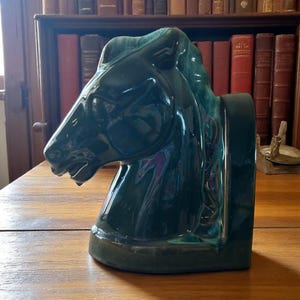 Puede incluir: Un sujetalibros de cerámica con forma de cabeza de caballo verde oscuro. El sujetalibros es brillante y tiene una melena y rasgos faciales detallados. Está sobre una superficie de madera, con una estantería al fondo.