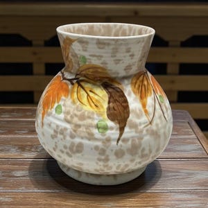 May include: A ceramic vase with a speckled cream and brown pattern. The vase features hand-painted leaves in shades of orange, yellow, and brown, with small green accents. The vase has a bulbous body and a flared rim, suitable for floral arrangements.