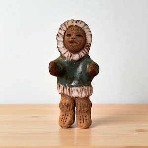 Puede incluir: Figura de cerámica de un inuit con un parka verde con capucha ribeteada de piel blanca. La figura tiene una cara sonriente y lleva botas marrones. La figura está sobre una superficie de madera.