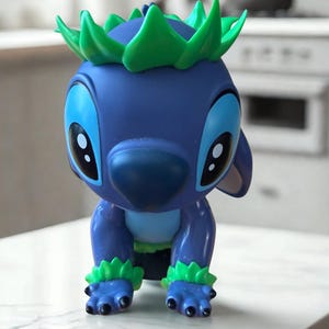 Puede incluir: Figura azul de un personaje de dibujos animados con ojos grandes, nariz grande y detalles verdes en la cabeza y los pies. La figura est&aacute; en posici&oacute;n agachada, con las manos y los pies en la superficie.