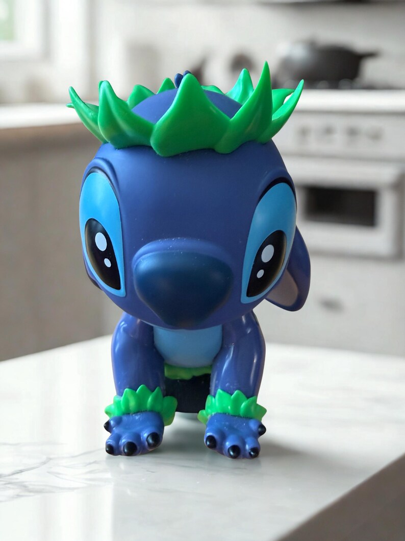Puede incluir: Figura azul de un personaje de dibujos animados con ojos grandes, nariz grande y detalles verdes en la cabeza y los pies. La figura est&aacute; en posici&oacute;n agachada, con las manos y los pies en la superficie.