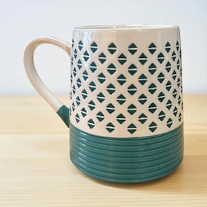 May include: A ceramic mug with a cream-colored upper section featuring a repeating pattern of teal diamonds. The lower portion is teal with horizontal ridges. The handle and the base of the handle are cream-colored with teal accents.