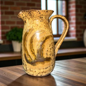 Puede incluir: Jarra de cerámica hecha a mano con una paleta de colores cálidos y terrosos. La jarra presenta una superficie moteada y texturizada con un asa prominente y un pico vertedor. El diseño incluye patrones arremolinados en tonos marrones y beige.