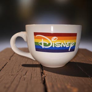May include: White ceramic mug with a large handle. The mug features the Disney logo over a rainbow-colored stripe design. The mug is sitting on a wooden surface.