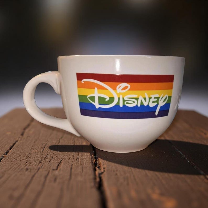 May include: White ceramic mug with a large handle. The mug features the Disney logo over a rainbow-colored stripe design. The mug is sitting on a wooden surface.