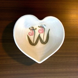 May include: Heart-shaped ceramic dish with a floral design. The dish is off-white with a painted design of two pink and brown flowers. The dish is sitting on a dark brown wooden surface.