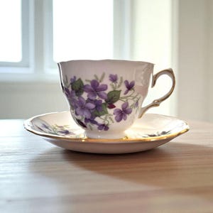 May include: A white teacup and saucer set with a floral violet design. The teacup has a gold handle and rim, and the saucer has a matching gold trim. The teacup and saucer are placed on a wooden surface.