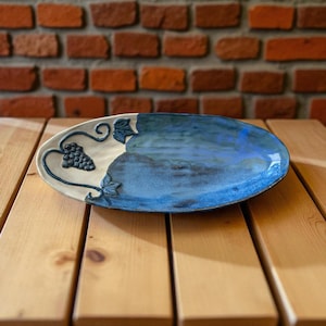 Könnte beinhalten: Ovale Servierplatte aus Keramik mit zweifarbigem Design. Die linke Seite ist cremefarben mit einem erhabenen Trauben- und Rankenmotiv. Die rechte Seite ist ein Farbverlauf in Blautönen. Die Platte steht auf einer Holzoberfläche.