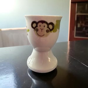 以下が含まれることがあります： 白いセラミックエッグカップ、猿の顔のデザイン。エッグカップは、茶色と緑のアクセントが付いた笑顔の猿の顔が特徴です。エッグカップは台座ベースとフレアトップを備えています。