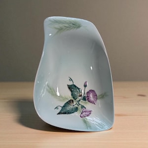 May include: A light blue, uniquely shaped ceramic dish. The dish is decorated with a floral design featuring green leaves and purple flowers. The design is painted on the inside of the dish, which has a smooth, glossy finish.