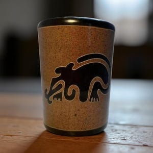 Puede incluir: Un vaso de chupito de cerámica marrón con un diseño de animal negro estilizado. El vaso tiene una textura moteada y un borde marrón oscuro. El diseño del animal es una silueta de una criatura con una cola larga y extremidades curvas. El vaso está sobre una superficie de madera.