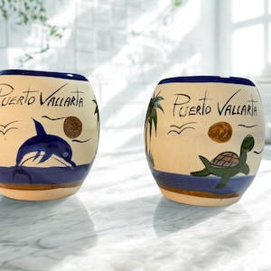 May include: Two ceramic, oval-shaped mugs with a cream base and blue accents. Each mug features the text "Puerto Vallarta" and a different ocean-themed illustration: a dolphin and a turtle. The mugs are approximately 10 cm tall.