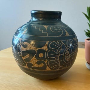 May include: A dark teal ceramic vase with a round body and a narrow neck. The vase features intricate, etched designs in a lighter tan colour, including floral and geometric patterns. The vase is sitting on a wooden surface.