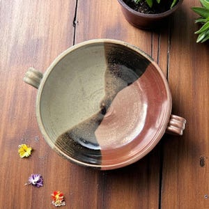 Peut inclure: Bol en céramique fait à la main avec un design tricolore unique. Le bol présente des sections vert olive, noir et brun rougeâtre. Il a deux petites poignées de chaque côté. Le bol est posé sur une surface en bois.