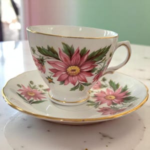 May include: White teacup and saucer set with a floral design. The cup and saucer feature pink flowers with yellow centers and green leaves. Gold trim accents the edges of both pieces, creating an elegant look.