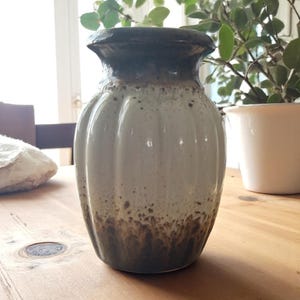 May include: A ceramic vase with a ribbed design. The vase has a dark brown rim and base, with a light gray body. The vase is sitting on a wooden table. A white plant pot is in the background.