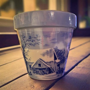 May include: A ceramic plant pot with a blue and white rural scene. The design features a barn, trees, and a cloudy sky. The pot has a light blue rim and a slightly tapered shape. The pot is sitting on a wooden surface.