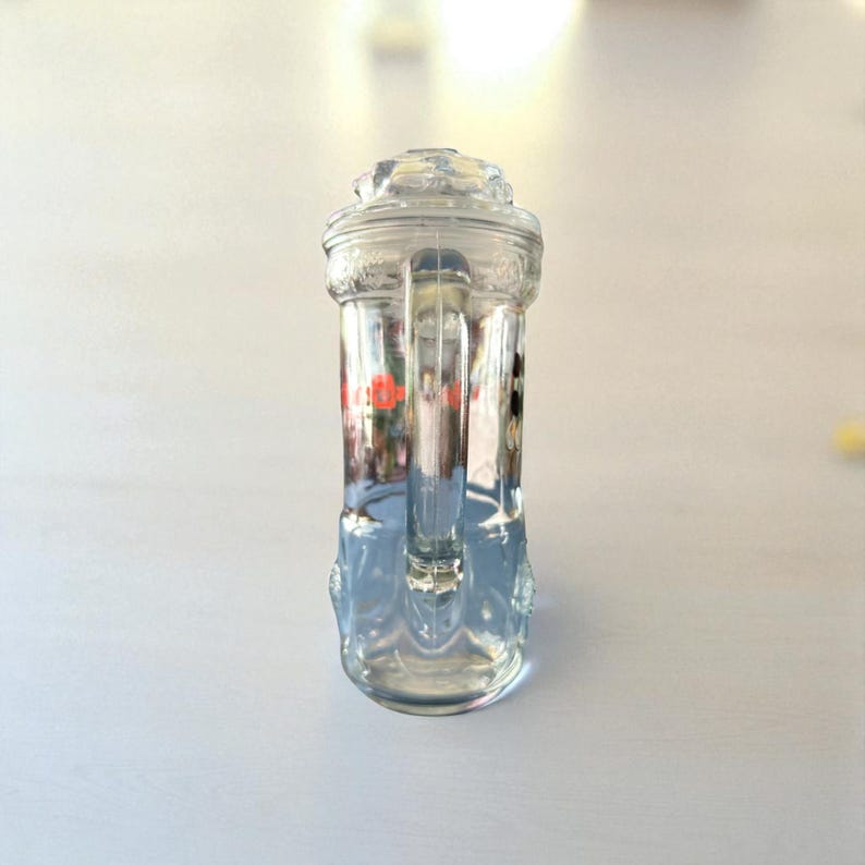 May include: A clear glass beer stein with a handle and a decorative lid. The stein has a cylindrical body with embossed details and a handle. The glass is transparent, allowing the contents to be visible. The stein is designed for holding beverages.