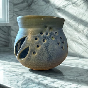 May include: A handcrafted ceramic essential oil diffuser with a unique design. The blue and brown vessel features multiple small holes and two larger cut-out shapes. The diffuser sits on a marble surface, with sunlight casting shadows.