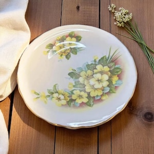 May include: A circular, cream-coloured ceramic plate with a gold edge. It displays a floral pattern of yellow flowers and green leaves. The plate rests on a wooden surface.