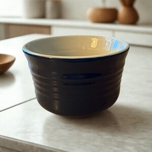 May include: A dark blue ceramic bowl with a cream-colored interior and a blue rim. The bowl has horizontal ridges around the exterior. The bowl is sitting on a white surface.