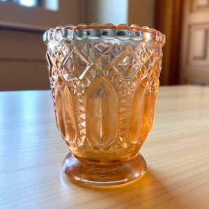 Puede incluir: Un portavelas de cristal teñido de naranja con un detallado patrón de diamantes y óvalos. El pequeño cristal decorativo tiene una base redonda y un borde festoneado. El portavelas es probablemente vintage y se encuentra sobre una superficie de madera.