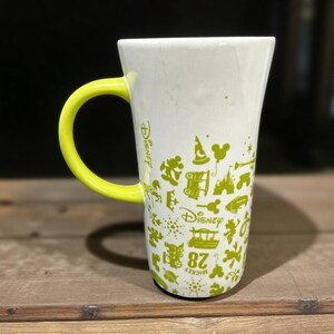 May include: White ceramic mug with a yellow handle. The mug features green Disney-themed graphics and text. The mug is on a wooden surface.