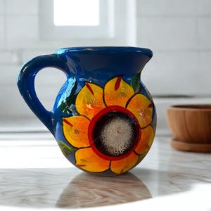 May include: A blue ceramic pitcher with a sunflower design. The pitcher has a large handle and a wide opening. The sunflower has yellow petals, an orange center, and a dark center. The pitcher is sitting on a marble surface.