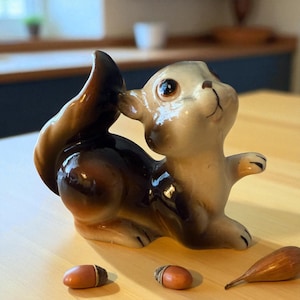 May include: A ceramic squirrel figurine in shades of brown, tan, and cream, posed with its tail curled up. The squirrel is holding its paw up to its mouth. Two acorns and a seed pod are in front of the figurine.