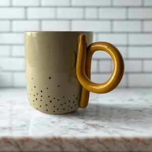 May include: A beige ceramic mug with a distinctive yellow handle. The mug has a series of small black dots on the lower section. The handle is a bright yellow loop. This handmade mug is suitable for tea, coffee, or other hot drinks.