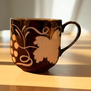 May include: A brown ceramic mug with a white grape and leaf design. The mug has a curved handle and a gold interior. The design features a cluster of grapes and a large leaf, with swirling vines. The mug is sitting on a wooden surface.