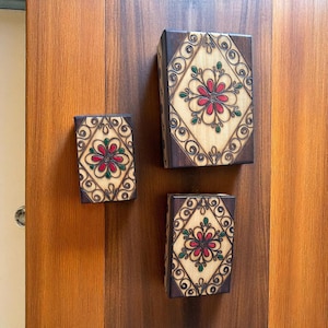 May include: Three wooden boxes with floral patterns. Each box has a central red flower with green and brown details, set in a diamond shape. The boxes are rectangular with dark brown borders and are likely hand-carved.