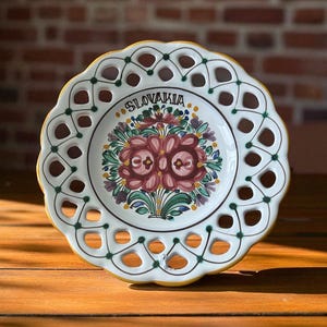 Puede incluir: Un plato de cerámica blanco con borde festoneado y calados decorativos. El plato presenta un diseño floral en tonos rosa, morado y verde, con la palabra "SLOVAKIA" en la parte superior. El borde está ribeteado en amarillo y verde.