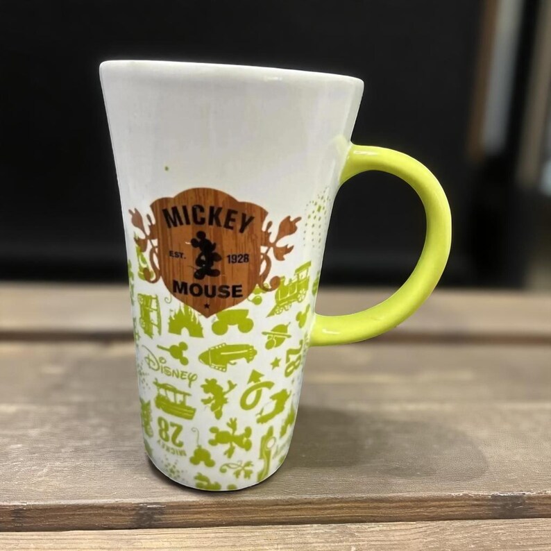 May include: White ceramic mug with a yellow handle. The mug features a brown and white Mickey Mouse logo and green Disney-themed graphics. The mug has a tall, tapered shape and is suitable for hot or cold beverages.