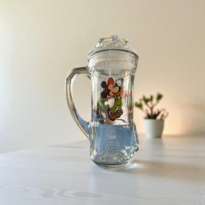 May include: Clear glass beer stein with a handle and a lid. The stein features a colourful image of Mickey Mouse in a red hat, green shirt, and brown shoes. The stein is sitting on a white surface.