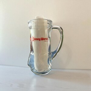 May include: Clear glass beer mug with a white lid and handle. The mug has the words "Disney World" in red lettering on the front. The mug is empty and sitting on a white surface.
