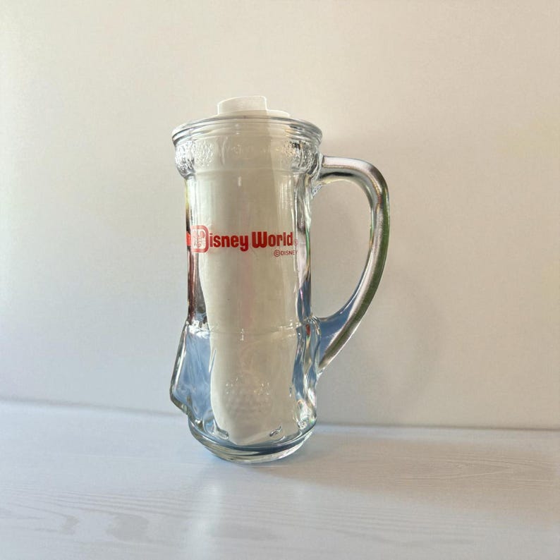 May include: Clear glass beer mug with a white lid and handle. The mug has the words "Disney World" in red lettering on the front. The mug is empty and sitting on a white surface.