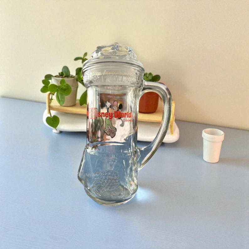 May include: Clear glass stein with a handle and lid, featuring a Mickey Mouse graphic and the text "Disney World" in red. The stein is empty and sits on a light blue surface. A small white cup is to the right.