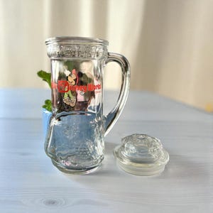 May include: Clear glass mug with a handle and a lid. The mug features a colorful graphic of Minnie Mouse and the words "Disney World". The lid is clear and round. The mug is sitting on a light blue surface.