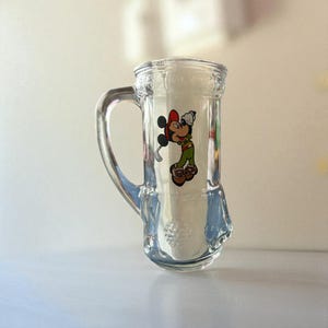 May include: Clear glass beer mug with a handle, featuring a colorful graphic of Mickey Mouse in a golf outfit. The mug has a textured design and a wide base. The Mickey Mouse graphic includes red, green, and brown colors.