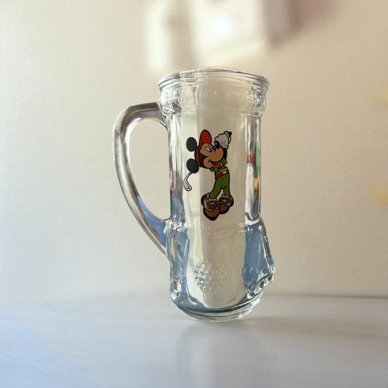 May include: Clear glass beer mug with a handle, featuring a colorful graphic of Mickey Mouse in a golf outfit. The mug has a textured design and a wide base. The Mickey Mouse graphic includes red, green, and brown colors.